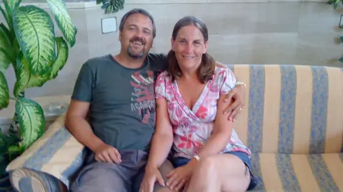 Caroline Rutley-Frayne Simon and Caroline sit on a stripey, yellow and blue sofa. Simon has brown hair and a small beard with flecks of grey. He is smiling. Caroline has brown hair in bunches and she is smiling at the camera too.