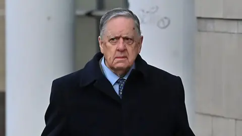 William Lloyd-Lavery walking outside. He is wearing a navy jacket over a light blue shirt with a navy tie with multi-coloured spots on it. He has grey hair. 