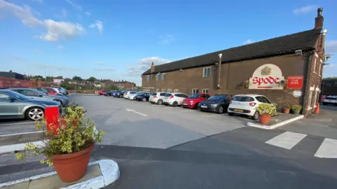 A car park at the Spode site in Stoke. There are a number of cars parked on both sides, with a pedestrian crossing in the foreground. There is a red Spode logo on the side of a building and plants in large pots separating sections of the car park.