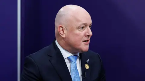 Side view of Christopher Luxon speaking. He's wearing a suit with a blue tie.