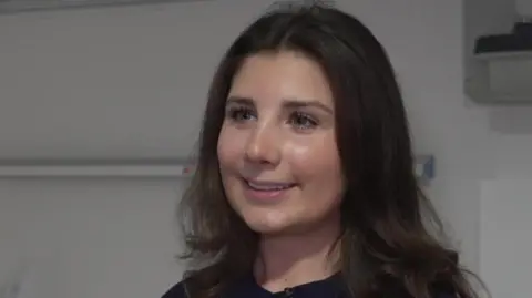 Lyra Cassell is standing in a hospital ward. Medical equipment is fastened to the wall in the background. She is smiling at the camera. 