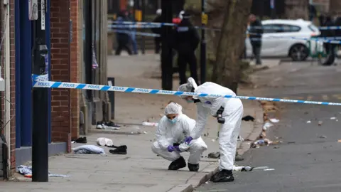 Darren Staples / AFP via Getty Images Forensic officers in white protective gear at the scene of an incident with a cordon in place in Friar Gate, Derby, on Sunday 29 March 2026. Debris remains on the road and cars are parked along the right in the background with out-of-focus police officers on the pavement.