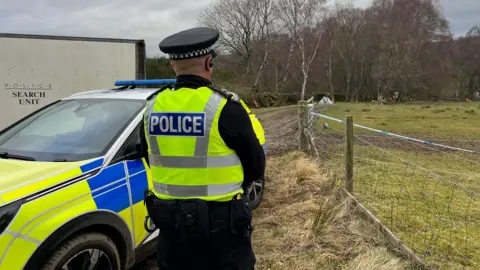 A police officer stands beside a police car guarding a taped-off incident on a muddy path near a field