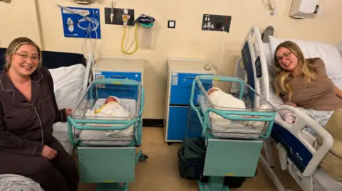 Abi Abi on the left and Jessica on the right with their babies in hospital cots in the middle. They are both smiling at the camera and in their hospital beds.