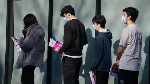 PA Media Four young people wearing face masks stand in a line outside a building with large glass panels, their shadows visible on the frosted glass behind them. Several are holding pink and purple NHS leaflets about meningitis and septicaemia. They are dressed in casual clothing including hoodies and jackets, standing quietly as they wait.