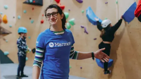 Kendal Wall Mel Adam is wearing a blue short-sleeved t-shirt under which she has a green, blue and white striped long-sleeved t-shirt. She appears to be mid conversation, with one arm out in an expressive manner. Behind her, and slightly out of focus, is a pale wood coloured climbing wall with holds or grips attached in a variety of colours. One child is climbing and another watches, both wearing helmets.