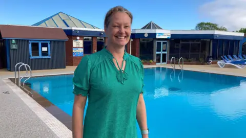 A woman wearing a green top standing next to an outdoor swimming pool. Two ladders lead into the pool. A building is behind her. The woman is smiling at the camera.