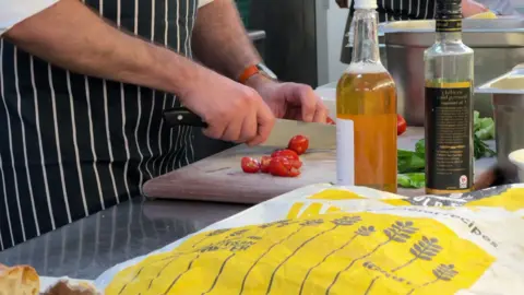 A chef in a kitchen cutting tomatoes on a wooden chopping board. There are other ingredients and bottles around him.