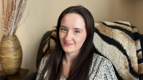 A photo of Freya sat down in front of a tiger print rug. She has long brown hair and is wearing a jumper. 