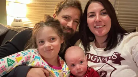 The photo shows Tom with his wife and children on a sofa. Tom has short brown hair and a well trimmed beard. He is wearing a black jumper. His wife has shoulder length dark brown hair. She is wearing a white jumper.