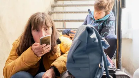 A mum wearing an orange puffer jacket looks anxiously at her mobile phone with a blue backpack beside her, while her young son sits on a metal staircase behind her, looking down.