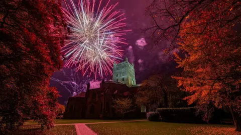 Jack Boskett Fireworks in front of Tewkesbury Abbey with orange and pink colours surrounding it.