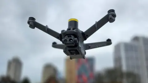 A drone in the air. The grey four-pronged equipment is in front of the blurred outline of tower blocks