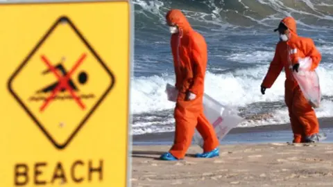 A beach closed sign is in the foreground while people in orange PPE walk on the sand in the backgound