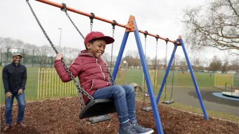Getty Images A young boy wearing a red baseball cap and red coat plays on the swings in a park playground, watched by his smiling father in a black jacket and brown cap.