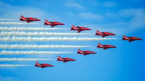 The RAF Red Arrows display team performing during an airshow, in an arrow formation against a blue sky