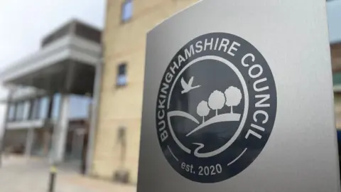 A view of a grey sign with the Buckinghamshire Council logo on it. A building can be seen behind it.