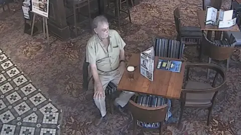 Crown Prosecution Service Photo taken from a ceiling mounted CCTV camera showing Jeremy Rickards wearing a beige shirt and trousers with a pint of beer in front of him as he sits at a table in  a pub with carpeted and tiled flooring. 