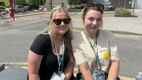 Grace McGrory/BBC Two women sat on a stone bench in front of a car park. They are both wearing a green and white lanyard. One woman has dark sunglasses on and blonde hair half tied up and the other has dark sunglasses on her forehead with her hair tied back. One is wearing a black top and the other a white top with a lemon on it