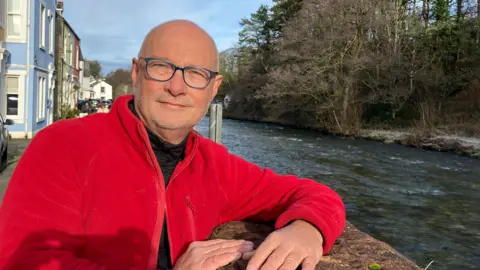BBC/Ed Hanson Jonty, who is bald and wearing blue glasses and a red fleece jacket, leans on a stone wall next to a river.