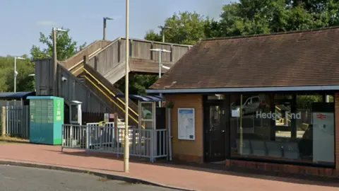Google Google Street View of Hedge End station entrance - a single-storey building with a pitched roof and a glass front. A stepped, timber-clad footbridge can be seen to the left crossing the railway line.