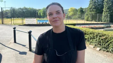 BBC/Charlotte Benton A woman standing on in front of grass tennis courts. She is smiling and wearing a black t-shirt and apron. The sky is blue in the background and tall evergreen trees stand along the side of the tennis court and behind it. 