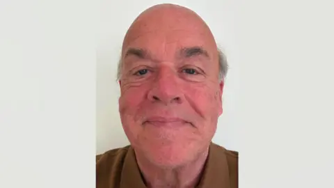 Graham Norwood A bald man with grey sideburns smiling in a selfie wearing a brown polo top.
