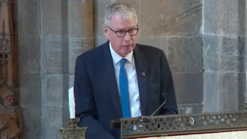 Dunblane Cathedral A man with short grey hair and glasses is wearing a black jacket, white shirt and blue tie as he stands at a church lectern
