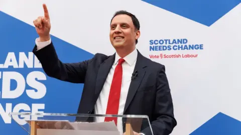 PA Media Anas Sarwar, who has short black hair, in front of a saltire background. He is wearing a business suit and a red tie, pointing in the air and smiling. 