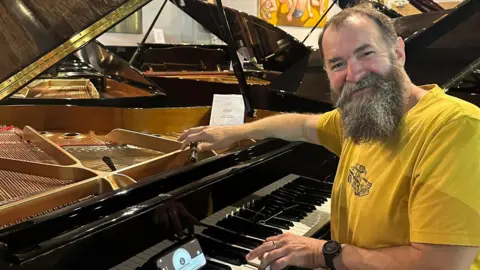 Simon Juniper Image of Simon. He is sat at a piano wearing a yellow t-shirt. He has a large beard.