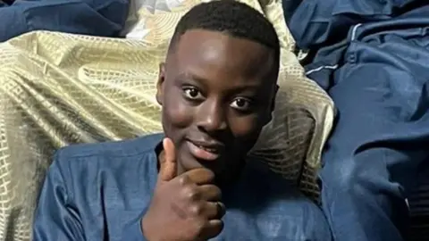 A smiling teenager sits and gives a thumbs up to the camera.