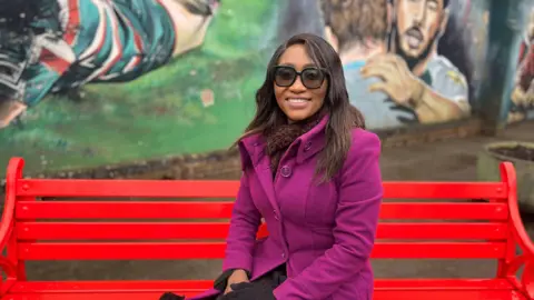 A woman sat on a bright red bench wearing a bright purple coat and black glasses