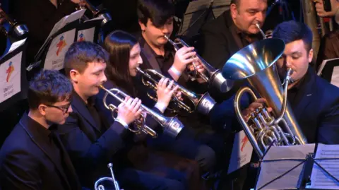 Jacob Hickson Five men and woman in black suits play brass instruments in a band.