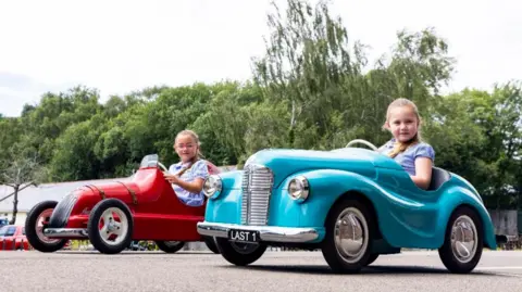 Austin Pedal Car Company A red Austin Pathfinder and a blue J40 side by side. Both are driven by young girls of around seven-years-old in blue summer dresses
