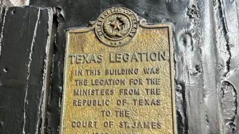 Close-up of a weathered metal plaque mounted on a textured surface, featuring the seal of the Republic of Texas and engraved text reading “Texas Legation,” explaining that ministers from the Republic of Texas were based there