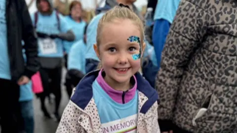 Elsie, wearing blue and purple face glitter, and a blue T-shirt as she finishes a charity run