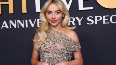 A headshot of Sabrina Carpenter wearing a glittery dress and smiling. She stands in front part of a sign. 