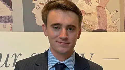 A brown haired man with a dark suit jacket, blue shirt and dark tie faces the camera, with a tapestry design in the background