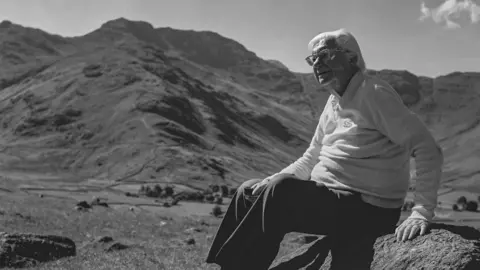 Chris Butterfield/Ken Shepherd Black-and-white photograph of Alfred Wainwright sitting on a rock with the fells behind him. He is looking to the left of the image, and is wearing a light jumper and dark trousers. He has white hair and has glasses.