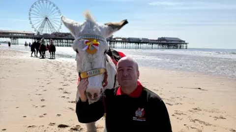 Mark poses on Blackpool beach with a white donkey named Alfie. Mark is bald and is smiling at the camera wearing a black company-branded jumper with a red polo shirt underneath. The pier and Big Wheel can be seen beyond the sand and sea in the background