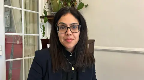 Woman wearing a dark blouse under a navy blazer. she has a gold pendant, long dark hair and black framed glassed. Behind her is a potted house plant and lightly coloured walls and a glass door on the left of the image.