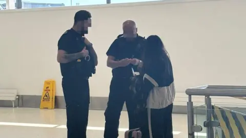 Two men in dark uniforms from Home Office Enforcement question a woman at an airport. Although both men's faces are blurred, one is tall with dark hair and a beard while the other is balding. The woman, who has long black hair, has her back turned to the camera. 