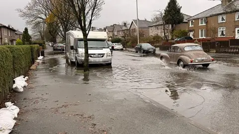 A motorhome is among a number of vehicles parked in a road flooded with water as a car is being driven through the surface water. Sandbags are seen on the left attempting to stop water flooding gardens.