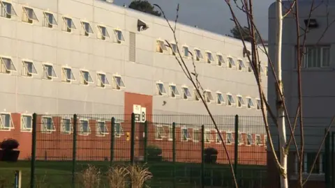 Parc Prison building from behind a fence. A tree is visible in the righthand corner, the building is a mix of red brick and grey - all windows are open. 