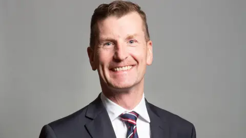 Dave Doogan, a smiling man wearing a suit with a stripy tie and side-parted blonde hair