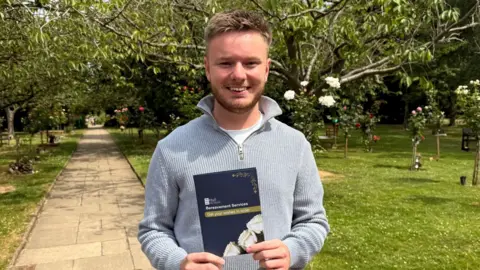 Councillor Ted Dolman standing on a path next to a lawned area. He is wearing a grey jumper and is holding a folder in front of him which says get your wishes in order on the cover.