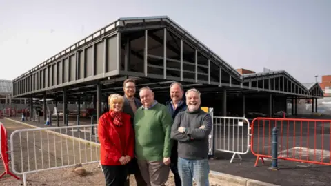 City of Wolverhampton Council Four men and a woman stand on the site of the market which is under construction behind them. The roof section is being built and barriers can be seen directly behind the group, who are all smiling at the camera.