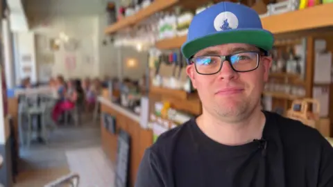 A picture of Gareth, who is wearing glasses and a cap, standing in front of a bar, there are customers in the background.