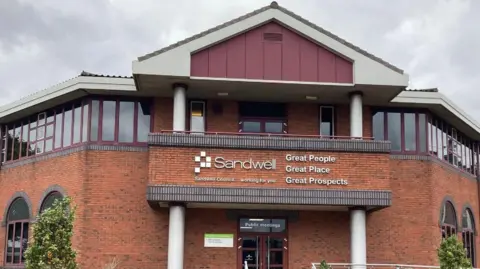 A redbrick two-storey building with a triangular roof. The building has windows all along the top floor, which have maroon window frames. The building says "Sandwell" in large white letters on the front.