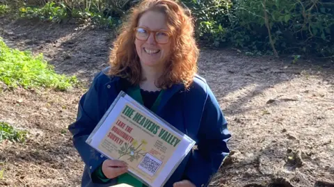 Emma Kernahan A woman with curly reddish hair, and glasses, wearing a blue jacket, is standing in a country lane, smiling to camera and holding an A4 piece of paper with information about the Heavens Valley on it.